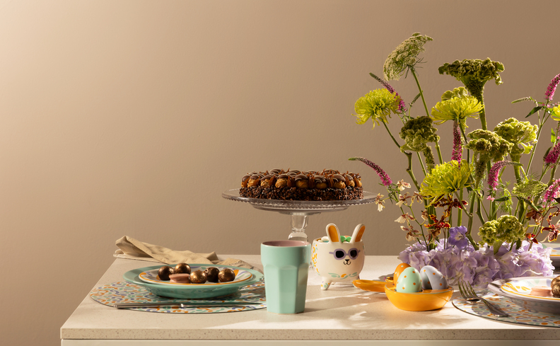 mesa de páscoa com receitas de doces, coelho de adorno decorativo, copo e vaso com planta, além de ovos de páscoa decorativo