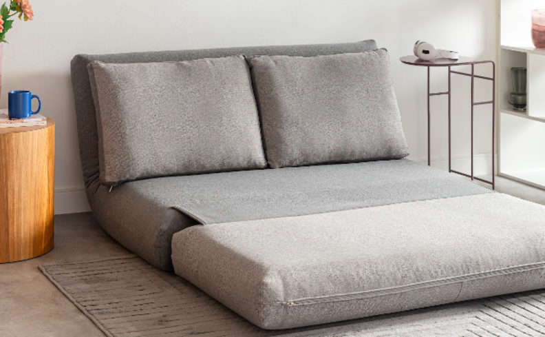 sofá cama para sala pequena na cor cinza com mesas de apoio uma de madeira e uma de aço, além de tapete e itens decorativos.