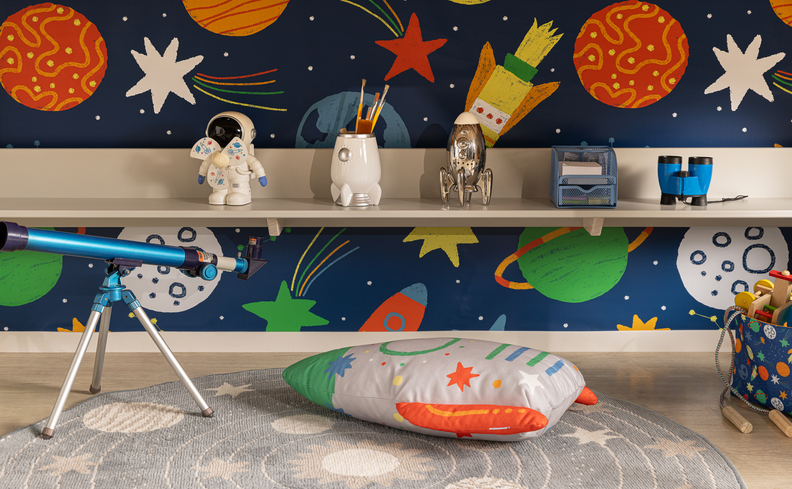 quarto infantil com parede de astronauta e galáxia, prateleira baixa com diversos itens decorativos, tapete, almofada e um microscópio.