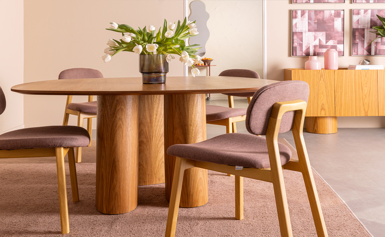 altura mesa de jantar em um modelo orgânico com a base no centro em madeira, além de quatro cadeira de madeira com o estafado na cor ruby rose e um vaso com flor no centro da mesa.