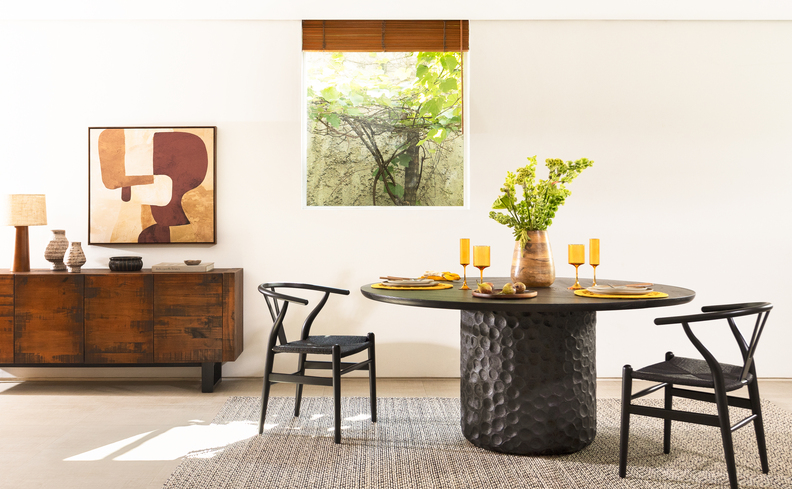 mesa de jantar oval com tampo de madeira e centro de madeira na cor preta, além de duas cadeiras pretas, vaso de flores na mesa, quadro e buffet aparador.