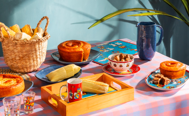 comida típica de festa junina com mesa posta completa com bolo, espiga de milho, vinho quente na caneca de cerâmica, vaso com planta e outros pratos típicos da época.