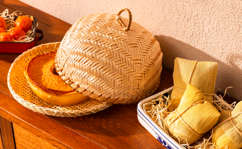 comidas de festa junina sobre uma mesa de madeira, com bolo de fubá em uma boleira com tampa de palha, pamonha e outros pratos típicos de São João.