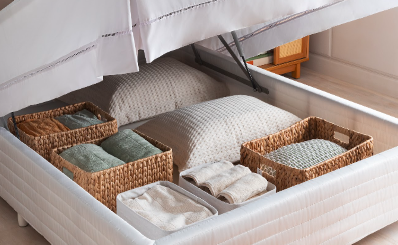 cama com baú organizado com caixas organizadoras de rattan, separando toalhas, travesseiros e jogos de cama pra otimizar o espaço.
