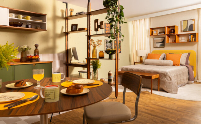 mesa para pequeno espaço em uma casa integrada com sala, cozinha e quarto, separado por uma estante vazada, além de cama, cadeiras, armários modulares, tapete e diversos itens decorativos.