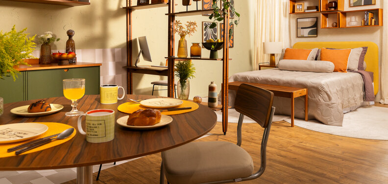 mesa para pequeno espaço em uma casa integrada com sala, cozinha e quarto, separado por uma estante vazada, além de cama, cadeiras, armários modulares, tapete e diversos itens decorativos.