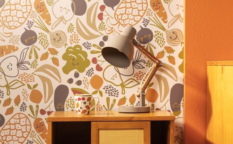 decoração de quarto infantil contendo: parede parte em papel adesivo colorido, parte em laranja; cômoda em madeira contendo uma xícara e uma luminária.