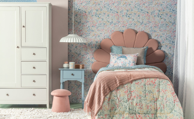 decoração de quarto infantil contendo: guarda-roupa branco; puffe rosa; mesa de cabeceira azul com luminária; cama com cabeceira rosa, jogo de cama colorido e edredom colorido; tapate branco.