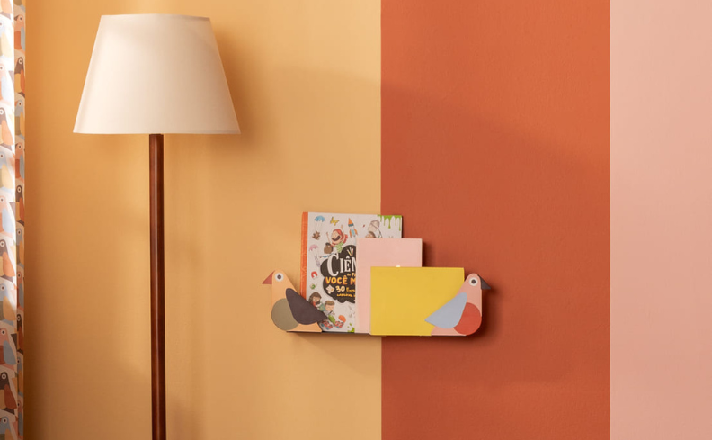 decoração de quarto infantil contendo: parede colorida dividida em laranja, vermelho e rosa; luminária de chão com cúpula branca; prateleira em forma de passarinho contendo livros.
