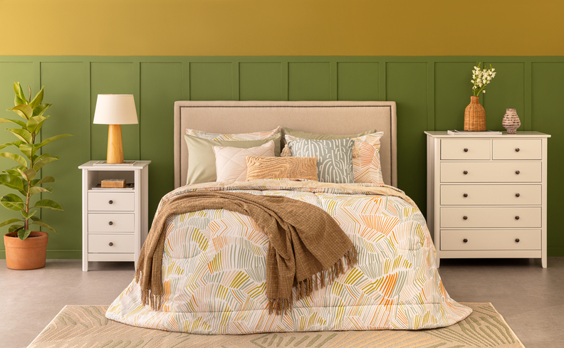 como organizar o quarto com mesa de cabeceira com gavetas, cama posta com roupa de cama em tons neutros, luminária de mesa, vaso com planta e diversas almofadas.