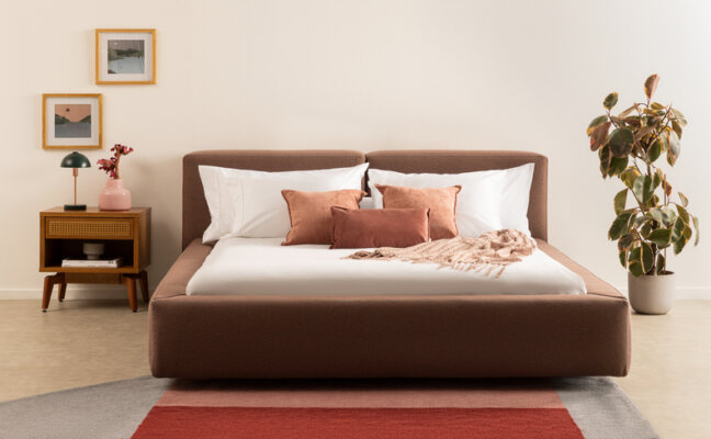 cor do ano em tons de marrom na cama, cabeceira e almofadas, além de tapete, mesa de cabeceira e vasos de planta