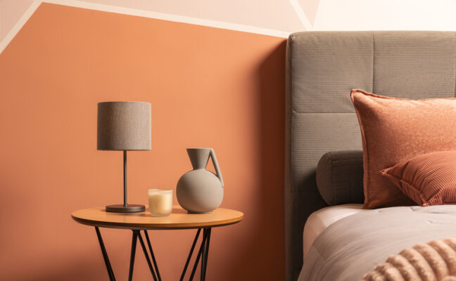 mesa de cabeceira para quarto com abajur, vaso e uma vela aromática em um ambiente com cama, roupa de cama e cabeceira