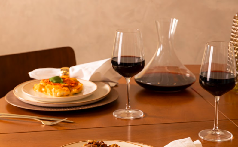 tipos de vinho tinto em uma mesa posta com taças, decanter de vinho, pratos e talheres com uma refeição para aproveitar o Dia dos Namorados