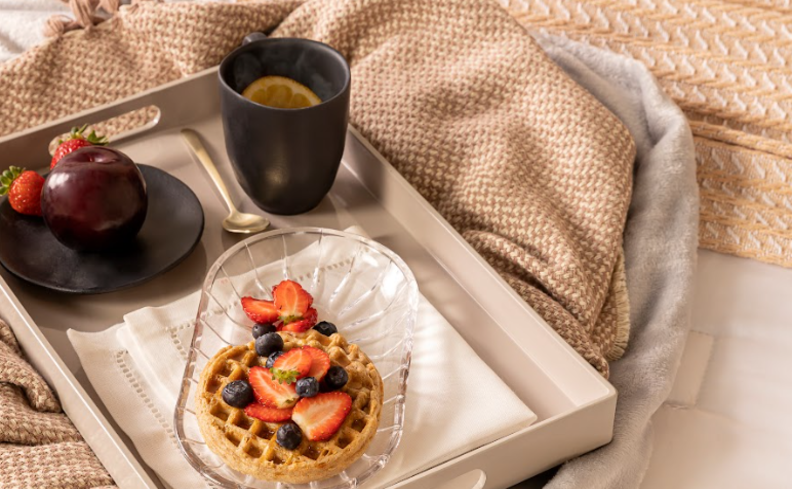 bandeja café da manhã na cama com prato com waffle e frutas, caneca com chá e um prato de sobremesa com frutas.