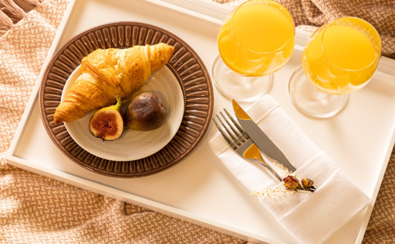 café da manhã na cama em uma bandeja com sousplat, prato de sobremesa, croissant, garfo e faca, além de duas taças com suco de laranja.