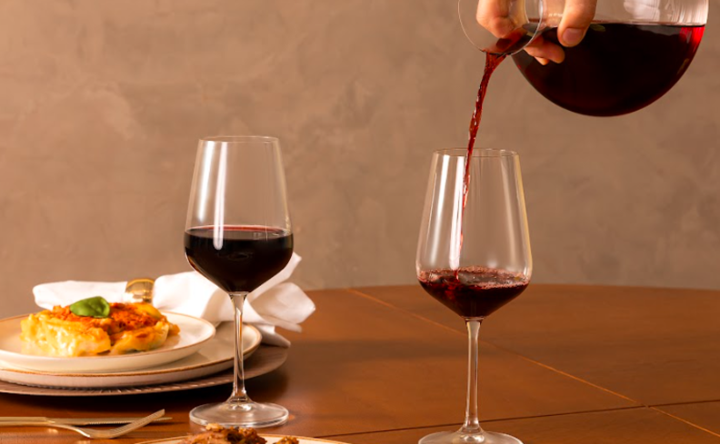 mesa posta com duas taças de vinho tinto e uma pessoa servindo vinho em um decanter