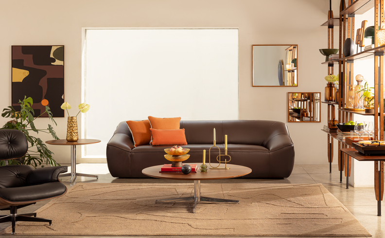 sala de estar com sofá em couro marrom, três almofadas coloridas laranjas de diversos formatos, estante com diversos itens decorativos, tapete e uma mesa de centro.
