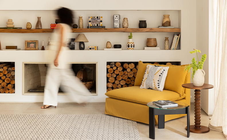 almofadas decorativas em uma poltrona amarela na sala de estar, com duas mesas de apoio, vaso decorativo e prateleiras com diversos itens de decoração.