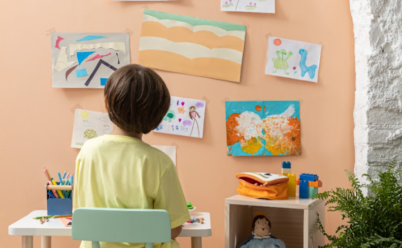 atividade de artes dia das crianças com um menino sentado em uma mesa infantil desenhando e com diversos desenhos pendurados na parede.