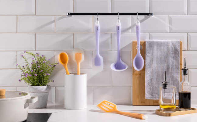 utensílios para cozinha de silicone em uma bancada completa com esses itens em roxo e laranja, além de tábua de corte e um vaso decorativo.