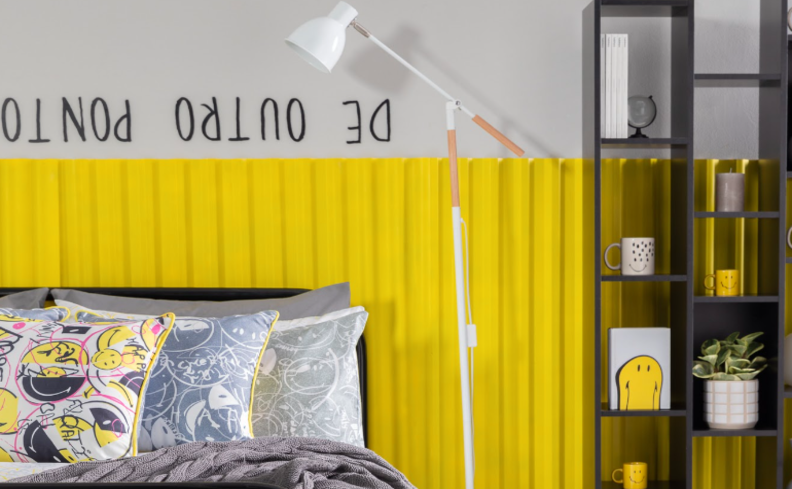 cor de quarto de adolescente com metade da parede amarela, cama com roupa de cama cinza com diversas almofadas e travesseiros, além de uma luminária de chão e uma estante com itens decorativos e livros.