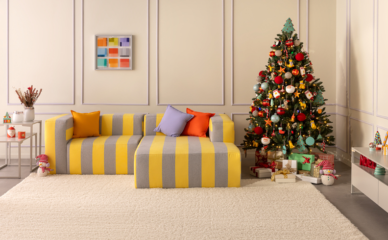 árvore de Natal pequena decorada na sala de estar com sofá colorido, tapete, almofadas, quadros decorativos e mesa lateral.