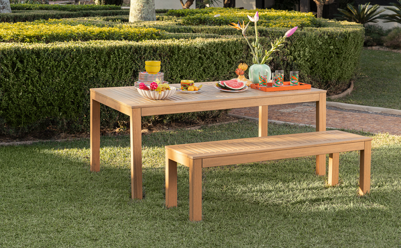 carnaval em casa com mesa posta de madeira e um banco, além de diversos pratos, copos, vaso com flores e comidas para refrescar no verão.