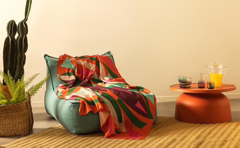 jogo de mesa posta carnaval para receber a família e amigos em um espaço descontraído com chaise de tecido, uma canga com cores vibrantes e uma mesa posta lateral com copos e taças.