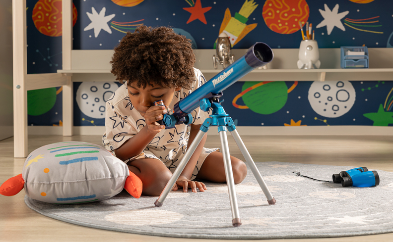 decoração astronauta no quarto infantil com tapete, almofada de foguete, parede com o tema via láctea e um menino sentado observando em um telescópio.