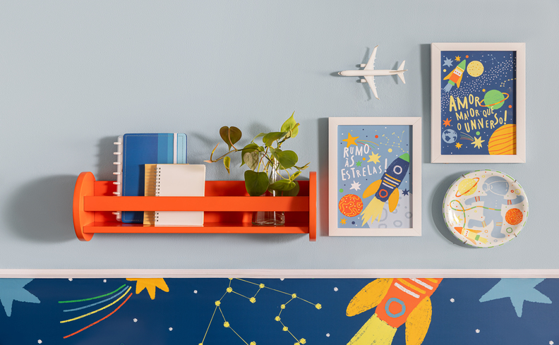 decoração para quarto infantil com parede no tema via láctea, prateleira laranja, quadros decorativos e diversos adornos de foguete e astronauta.