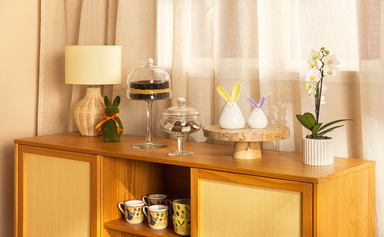 decoração de Páscoa para casa com um buffet com luminária de mesa, canecas, boleira, adornos decorativos de Páscoa e vaso com planta.