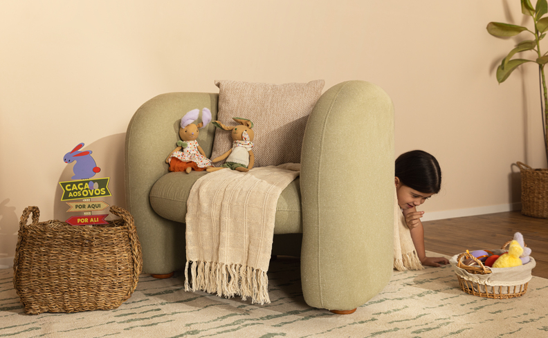 ideias para Páscoa para divertir toda a família com caça aos ovos com uma criança escondida atrás de uma poltrona na sala de estar com vaso, manta para sofá e coelhos decorativos.