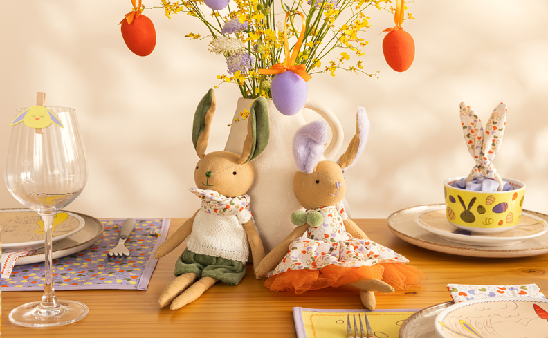 mesa de Páscoa com vaso decorativo com ovos pendurados em plantas artificiais, bonecos de coelho, pratos, tigelas, talheres e jogos americanos temático de coelho.