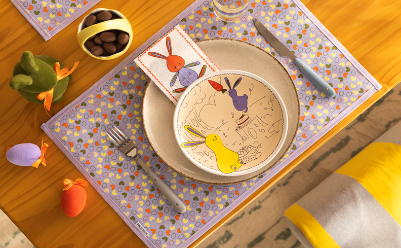 mesa posta Páscoa com jogo americano com tema de coelhos, pratos, cestinho com ovos de chocolate, talheres, guardanapos e pratos.
