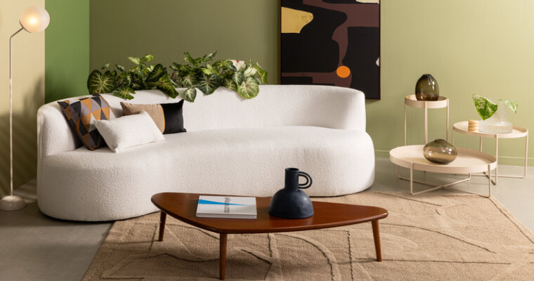 sofá de canto para sala pequena, modelo branco curvo, além de mesa de centro, três mesas de apoio com tamanhos diferentes, quadro decorativo, tapete e diversos vasos com plantas.