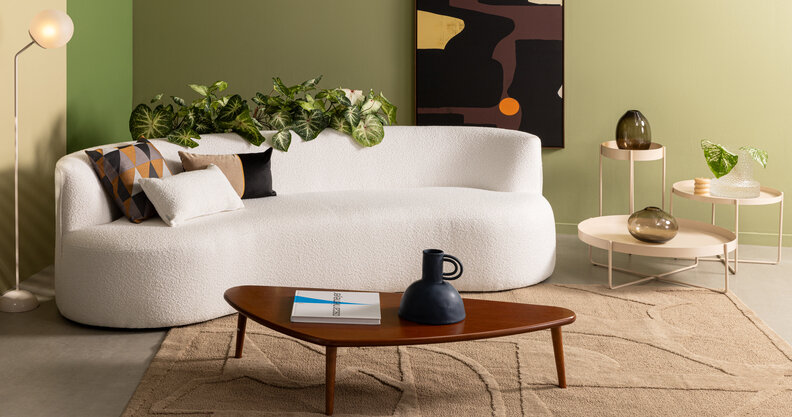 sofá de canto para sala pequena, modelo branco curvo, além de mesa de centro, três mesas de apoio com tamanhos diferentes, quadro decorativo, tapete e diversos vasos com plantas.