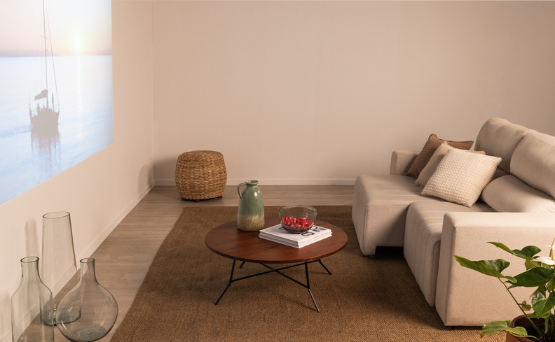 sofá retrátil branco para sala de estar pequena, mesa de centro redonda, um puffe no canto da sala, vaso com planta e um projetor na parede.
