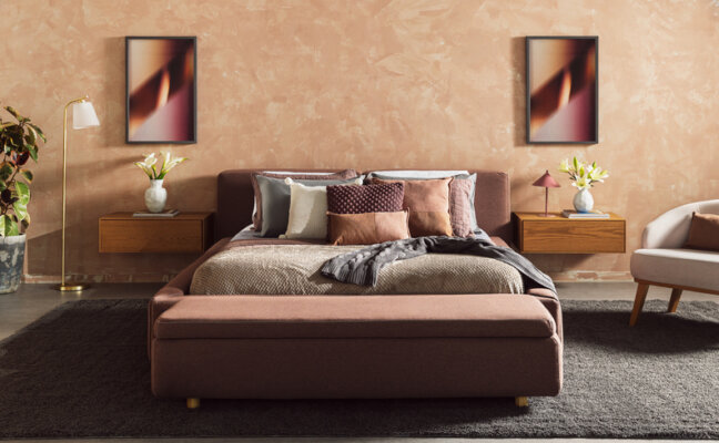 quarto com cama posta em tons de cinza e marrom e rosa, quadros decorativos, mesa de cabeceira, poltrona, vasos com plantas, roupa de cama completa e diversos travesseiros e almofadas.