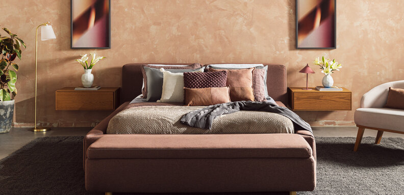 quarto com cama posta em tons de cinza e marrom e rosa, quadros decorativos, mesa de cabeceira, poltrona, vasos com plantas, roupa de cama completa e diversos travesseiros e almofadas.