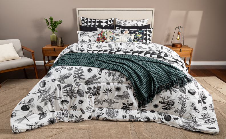 cama posta cinza, branca e verde, com peseira, edredom, travesseiros, almofadas, mesas de cabeceira, vasos cm plantas e luminária de mesa. Além de poltrona e cabeceira para decorar o quarto.