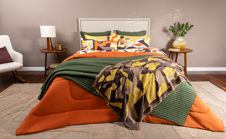 cama posta colorida com edredom, peseira, almofadas e travesseiros nas cores verde, laranja e marrom. Além de mesas de cabeceira, abajur, vaso com planta e poltrona.