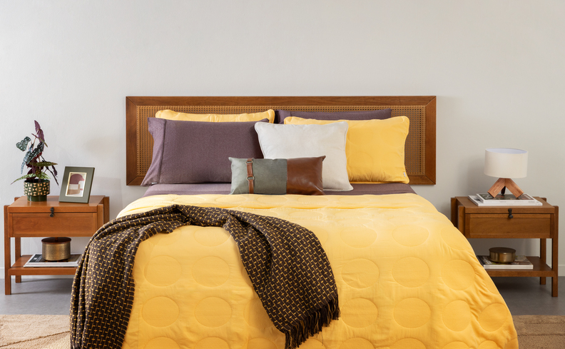 cama posta simples em um quarto com cama, cabeceira, roupas de cama em amarelo e marrom, além de diversas almofadas, travesseiros, mesa de cabeceira, vasos decorativos com plantas e abajur.
