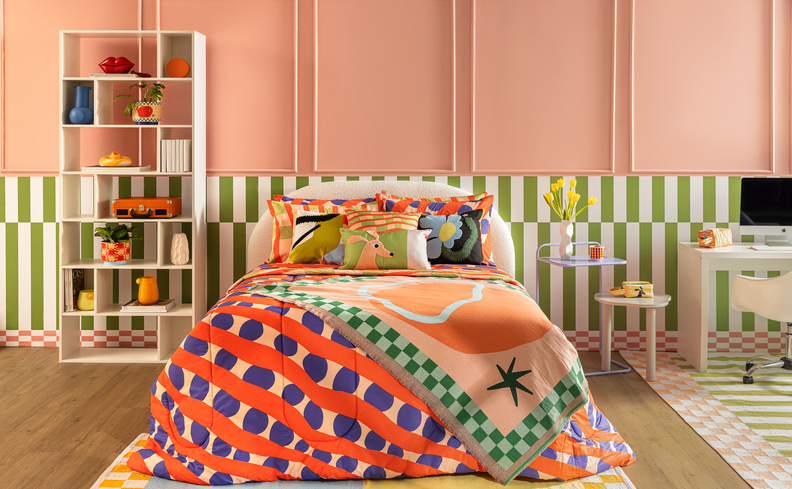 dicas de como arrumar cama em um quarto com cama, roupas de cama coloridas com edredom, almofadas e diversos travesseiros, além de tapete, estante, mesa de cabeceira, escrivaninha, cadeira de escritório e diversos itens decorativos.