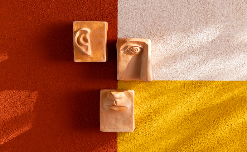 três peças decorativas de artesanato brasileiro penduradas na parede, uma orelha, um olho e uma boca escupidas à mão.