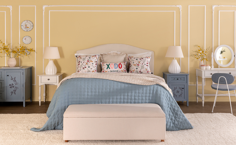 ideias de cama posta em um quarto com cama com roupas de cama em azul e branco, diversos travesseiros e almofadas, banco baú no pé da cama, mesas de cabeceira brancas com abajur, cômoda azul, penteadeira e uma cadeira.