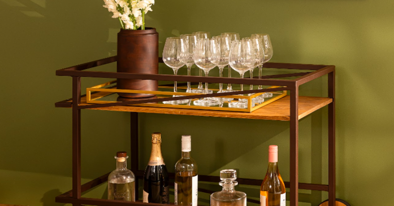 sala de estar com carrinho de bar de madeira com duas prateleiras com diversas taças, vaso com flores e várias garrafas de bebidas.