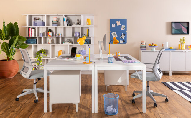 ambiente com móveis para home office com duas escrivaninhas brancas, cadeiras ergonômicas, estante branca, tapete, vaso com planta e itens decorativos para compor o escritório compartilhado para o casal.