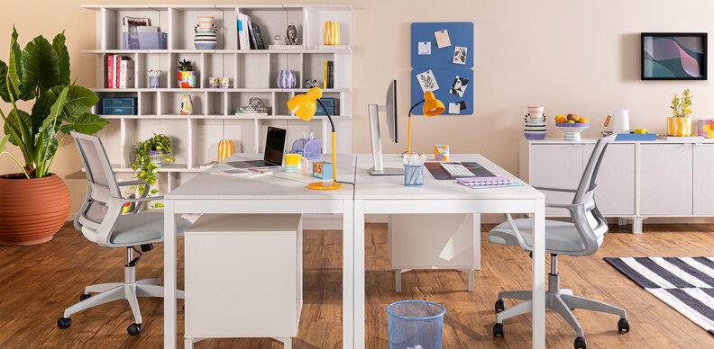 ambiente com móveis para home office com duas escrivaninhas brancas, cadeiras ergonômicas, estante branca, tapete, vaso com planta e itens decorativos para compor o escritório compartilhado para o casal.