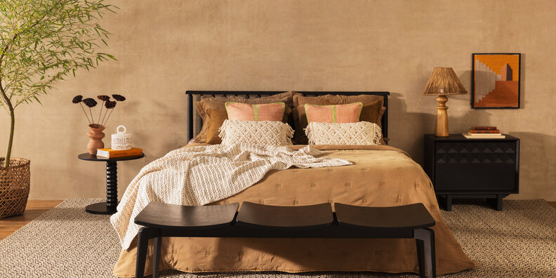 decoração boho em casa no quarto com tons neutros e terrosos com cama, peseira, roupa de cama, almofadas e travesseiros. Além de tapete, mesas de cabeceira, luminária de mesa e quadros decorativos e vasos com plantas.