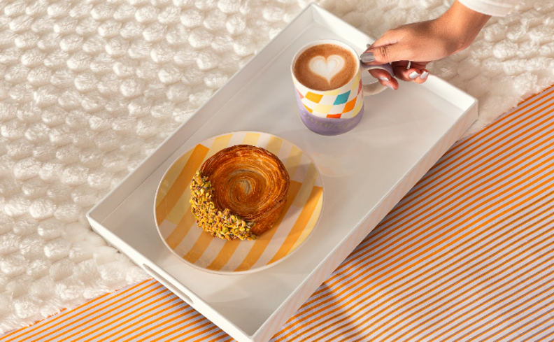 canecas de porcelana com estampa colorida em formatos geométricos em uma bandeja com um cinnamon roll e a caneca com café com um coração na espuma do leite.